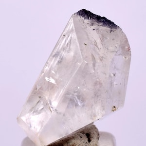 Puede incluir: Un espécimen de cuarzo cristalino transparente con una punta morada oscura. El cuarzo está facetado, con una base natural y rugosa. El cristal se presenta sobre un fondo neutro, resaltando su claridad y forma.