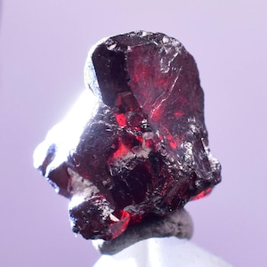 May include: A raw, dark red garnet gemstone with a rough, textured surface. The gemstone has a crystalline structure and displays areas of deep red color. The garnet is set against a soft, light purple background.