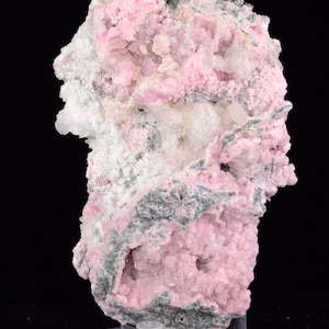 May include: A close-up of a mineral specimen with a mix of colours. The dominant colour is a soft pink, with areas of white and grey. The mineral has a textured, crystalline appearance, and is displayed on a clear stand.