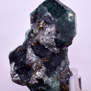 May include: A dark green mineral specimen featuring a large, faceted crystal formation. The mineral has a metallic, silver-grey matrix with small, golden inclusions. The specimen is displayed on a clear stand against a white background.
