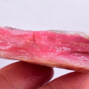 May include: A raw, pink rhodochrosite gemstone with a layered appearance. The stone displays a vibrant, deep pink color with lighter pink and beige inclusions. The rough, natural texture of the stone is visible.