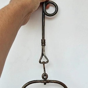 May include: Antique metal oil lamp with a hook and handle. The lamp has a circular base and a small rooster-shaped detail. The metal has a dark, aged appearance, suggesting it is vintage.