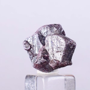 May include: A cluster of small, metallic, purple-grey crystals with a silvery sheen. The crystals are arranged in a cluster and have a rough, irregular surface.