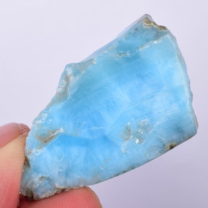 Puede incluir: Una pieza de Larimar en bruto, translúcida y de color azul claro. La piedra tiene un aspecto nublado y veteado con vetas blancas y grises.