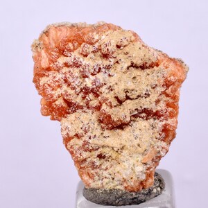 May include: A cluster of orange-red crystals with a rough, textured surface. The crystals are set against a white background.