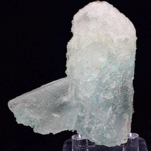 Może przedstawiać: Jasnoniebieski okaz minerału o krystalicznej strukturze. Minerał ma matowy wygląd z obszarami jasnoniebieskiego i białego. Okaz jest zamontowany na przezroczystym stojaku na czarnym tle.