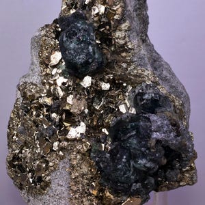 May include: A close-up of a mineral specimen with a matrix of sparkling, golden pyrite crystals. Dark green, crystalline formations are embedded within the matrix, creating a striking contrast. The overall texture is rough and uneven.