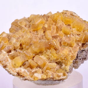 Puede incluir: Primer plano de un espécimen mineral que presenta un grupo de estructuras cristalinas de color amarillo dorado. Los cristales están agrupados sobre una matriz de color más claro, con un toque de gris en la base. El aspecto general es rugoso y texturizado.