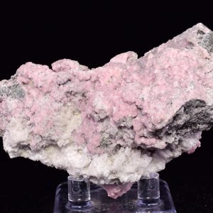 May include: A mineral specimen with a mix of pink, white, and grey crystalline structures. The pink crystals are the most visible, with a granular texture. The specimen is displayed on a clear stand against a black backdrop, accentuating its natural form.