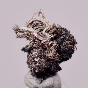 Puede incluir: Primer plano de un espécimen mineral plateado. El espécimen presenta una estructura compleja y texturizada con áreas plateadas y oscuras, posiblemente oxidadas. Se asienta sobre una base redondeada de color claro, sobre un fondo suave y neutro.