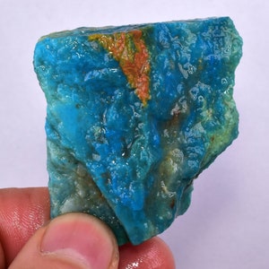 Może przedstawiać: Surowy, niebiesko-zielony kamień szlachetny z jasno pomarańczową żyłą przechodzącą przez niego. Kamień ma gładką, wypolerowaną powierzchnię i jest naturalnym, surowym okazem.