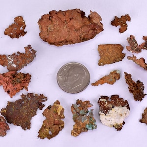 Puede incluir: Una colección de piezas de cobre de color marrón rojizo de forma irregular, algunas con toques de verde y blanco, dispersas sobre una superficie blanca. Se incluye una moneda de 10 céntimos para referencia de tamaño, mostrando los diferentes tamaños de los fragmentos de cobre.