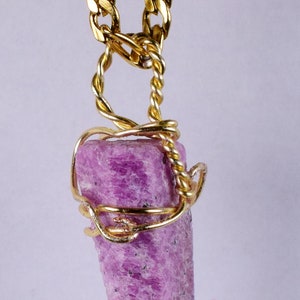 Puede incluir: Un colgante de piedra preciosa rosa envuelto en alambre dorado, colgando de una cadena de oro.