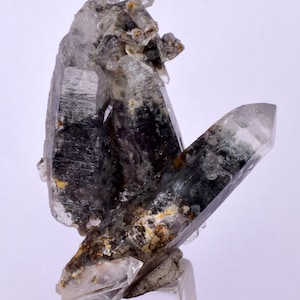 Puede incluir: Un grupo de cristales de cuarzo transparentes con inclusiones negras y verdes. Los cristales están dispuestos en una formación vertical, con el cristal más grande en la parte superior. Los cristales son translúcidos y tienen un brillo vítreo.
