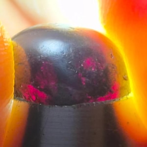 Puede incluir: Una piedra preciosa oscura, lisa y redondeada con una superficie pulida. Cuando se retroilumina, la piedra revela inclusiones internas de color magenta y rojo intenso que brillan con fuerza contra el exterior oscuro y opaco del mineral.