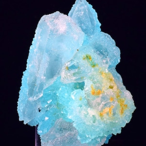 Może przedstawiać: Zbliżenie okazu mineralnego z skupiskiem jasno niebieskich kryształów. Kryształy mają krystaliczną strukturę i półprzezroczystą jakość, z żółtymi i zielonymi wtrąceniami. Tło jest czarne.