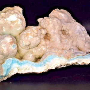 May include: A natural mineral specimen with a layered structure. The piece features a light beige color with areas of pale blue and green. The overall shape is irregular and organic, with a textured surface.