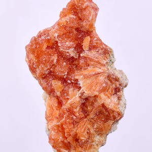 Op de afbeelding: Een ruw mineraal specimen, voornamelijk oranje van kleur, met een kristallijne structuur. Het oppervlak is ruw en getextureerd, met verschillende tinten oranje. De vorm is onregelmatig en gekarteld, met enkele doorschijnende gebieden.