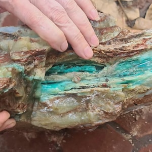 Może przedstawiać: Obróbka kamienia z warstwami brązu, zieleni i turkusu. Kamień jest trzymany w dłoniach, ukazując naturalną fakturę i wariacje kolorystyczne. Turkus jest dominujący w centrum.