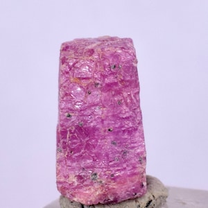 Puede incluir: Un cristal de rubí rosa vibrante con una textura rugosa y natural. El cristal tiene forma de prisma rectangular con una parte superior ligeramente puntiaguda.