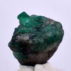 May include: A rough, green emerald gemstone with a dark green and black matrix. The emerald has a pointed, crystal-like shape.