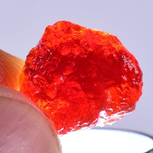 Könnte beinhalten: Ein leuchtend roter, durchscheinender, unregelmäßig geformter Edelstein. Der Edelstein hat eine raue, strukturierte Oberfläche.
