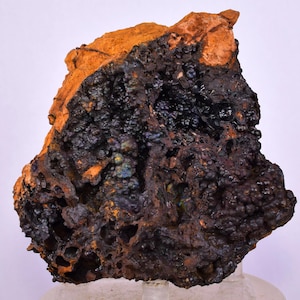 Puede incluir: Una roca áspera e irregular con una superficie oscura y texturizada. La roca presenta una mezcla de colores, incluyendo marrón oscuro, negro y naranja. Algunas áreas tienen una apariencia burbujeante, mientras que otras son más sólidas. Probablemente un espécimen mineral.