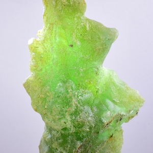 May include: A large, green, translucent, irregularly shaped piece of natural glass, known as Libyan Desert Glass, on a clear display stand.