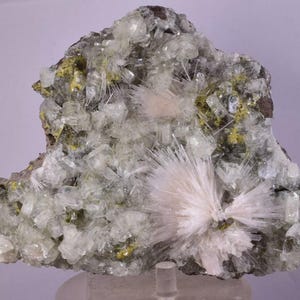 Może przedstawiać: Naturalny okaz minerału z przezroczystymi, krystalicznymi strukturami i jasnozielonym i żółtym odcieniem. Okaz ma szorstką, teksturowaną powierzchnię i centralną, promienistą formację białych, igiełkowatych kryształów.