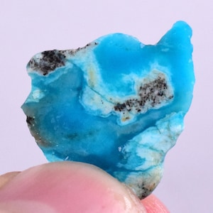 Puede incluir: Una pieza de turquesa áspera e irregular con un patrón azul y blanco e inclusiones negras. La turquesa es de color azul claro y tiene una superficie lisa y pulida.