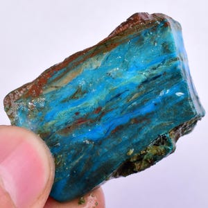 Puede incluir: Una pieza natural y rugosa de ópalo azul y verde con vetas marrones y blancas. El ópalo es translúcido y tiene una superficie lisa y pulida.