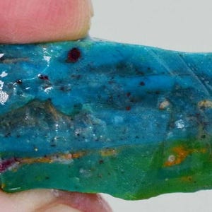 Puede incluir: Una gema en bruto vibrante con un gradiente de color llamativo. La piedra transita de un verde azulado profundo a un turquesa brillante, con toques de verde y marrón. La superficie tiene una textura natural y rugosa.
