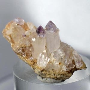 Puede incluir: Un grupo de cristales de cuarzo con puntas de amatista. Los cristales son de un blanco translúcido y las puntas de amatista son de un púrpura intenso. La base es una roca rugosa de color marrón claro. Los cristales están dispuestos en una formación natural y orgánica.
