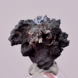 May include: A close-up of a mineral specimen. It features a dark, textured matrix with delicate, silver-coloured wire-like structures. The mineral has a rough, uneven surface and sits on a light-coloured base, with a soft, blurred background.