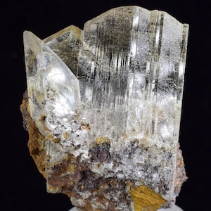 May include: A cluster of clear, glassy, elongated crystals on a brown, rocky matrix. The crystals have a striated texture and are translucent.
