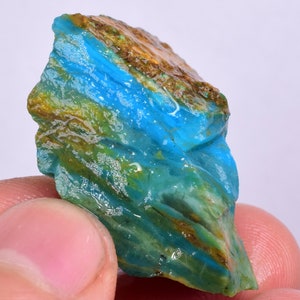 Może przedstawiać: Surowy, naturalny kawałek opalu w kolorze niebieskim i zielonym z brązowymi i białymi inkluzjami. Opal jest półprzezroczysty i ma warstwową strukturę.