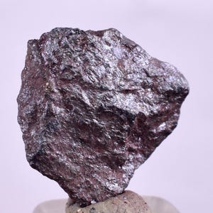 May include: A rough, dark gray, metallic rock with a slightly iridescent sheen. The rock has a jagged, irregular shape.