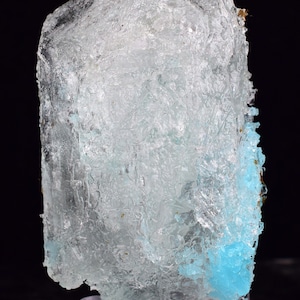 Może przedstawiać: Przezroczysty, krystaliczny okaz minerału o jasnoniebieskim odcieniu. Minerał ma szorstką, teksturowaną powierzchnię i mały obszar o żywym turkusowym kolorze. Okaz jest prezentowany na czarnym tle.