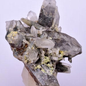 Puede incluir: Un grupo de cristales de cuarzo ahumado con una matriz mineral verde. Los cristales son transparentes y tienen un ligero tono ahumado. La matriz es verde y tiene una textura rugosa.