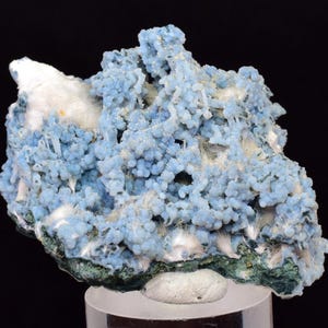 May include: A close-up of a mineral specimen featuring a cluster of small, round, light blue formations. The mineral has a textured appearance with white and green accents, set against a black background. The specimen is mounted on a clear stand.