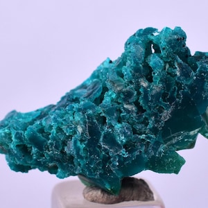 Puede incluir: Un espécimen mineral de color verde azulado vibrante con una estructura cristalina. El mineral tiene una superficie rugosa y texturizada y está montado sobre una pequeña base transparente. El color es un verde azulado profundo y saturado.