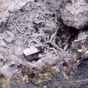 May include: A close-up of a dark gray, metallic mineral crystal with a sharp, geometric shape. The crystal is embedded in a white, rocky matrix with smaller, red crystals scattered throughout.