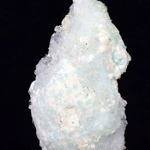 Może przedstawiać: Zbliżenie okazu mineralnego w kolorze jasnoniebieskim i białym. Minerał ma krystaliczną strukturę i szorstką, teksturowaną powierzchnię. Tło jest czarne.