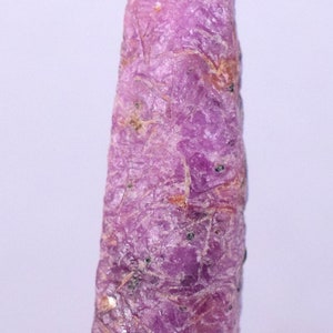 May include: A pink ruby gemstone with a rough, natural finish. The ruby is a long, thin, and pointed shape.