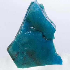 Puede incluir: Un vibrante espécimen mineral de color turquesa y forma irregular. La piedra presenta una estructura cristalina con áreas de azul más oscuro y toques de rojo. La superficie refleja la luz, mostrando su textura natural y variaciones de color. Sobre fondo blanco.