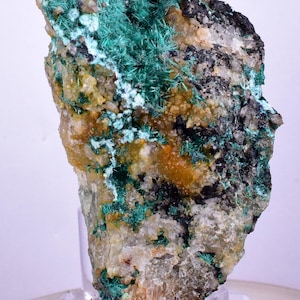 May include: A natural mineral specimen featuring a matrix of various colors. The rock displays a mix of green, black, orange, and white hues. The green formations appear as needle-like crystals. The mineral is mounted on a clear stand.