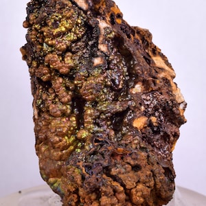 Könnte beinhalten: Ein großer, unregelmäßig geformter Stein mit einer rauen, strukturierten Oberfläche. Der Stein ist mit einer Schicht aus irisierendem, metallisch aussehendem Material bedeckt, das aus einer Mischung aus Grün, Gold und Braun zu bestehen scheint. Der Stein liegt auf einer weißen Oberfläche.