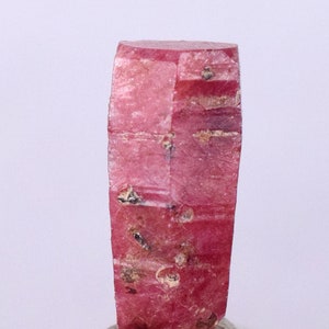 May include: A deep red ruby crystal with a faceted, elongated shape. The crystal has a rough, natural texture and is displayed on a grey base.