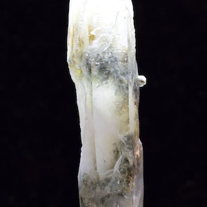 Puede incluir: Un cristal de cuarzo transparente con inclusiones de minerales verdes y marrones. El cristal es puntiagudo en la parte superior y tiene una base rugosa y natural.