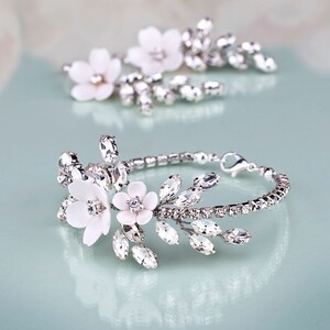 K&ouml;nnte beinhalten: Ein silbernes Armband und ein passendes Haaraccessoire, beide mit wei&szlig;en Blumenakzenten und funkelnden Kristallen verziert. Das Armband hat ein filigranes Kettendesign, ideal f&uuml;r Hochzeiten. Das Wort "GRACIA" ist sichtbar.